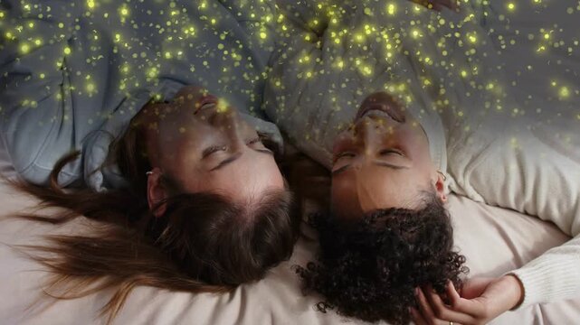 Two women lying on bed, sparkles drifting above initiating soft gaze and hair-touching for health