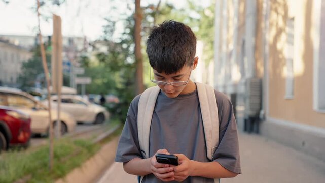 School student with backpack and glasses checking smartphone while walking in city street. Authentic education, real people and daily routine concept.