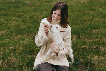 Naklejka premium A woman in a chunky beige sweater holds a white goat kid in a grassy field, sharing a tender moment of affection outdoors, showcasing rural lifestyle, animal companionship, and simple farm life.
