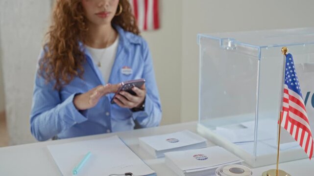Woman tapping smartphone with finger, voted sticker on shirt beside clear ballot box and american flag in polling building; pride duty.