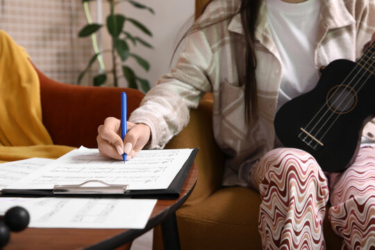Beautiful young female composer writing notes while playing ukulele on armchair in music studio, closeup