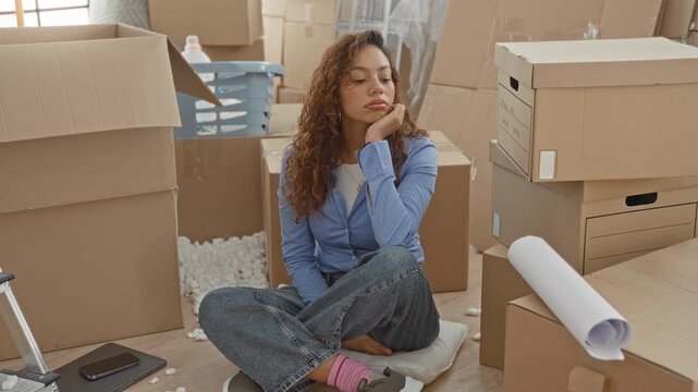 Woman seated cross legged with hand on cheek among cardboard moving boxes in building; pensive reflection.