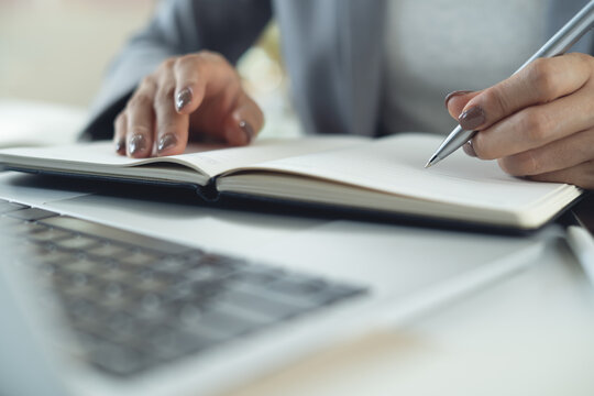 Business woman writing in notebook at office with laptop on desk, business planning, doing online research, close up