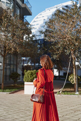 Fototapeta premium Young woman with red hair wears a long red dress and carries a brown leather bag while walking outdoors in a sunny urban park with autumn trees around her.