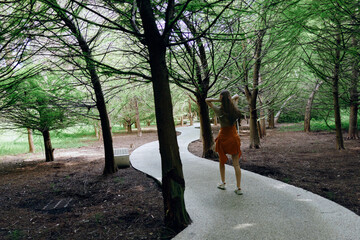 Fototapeta premium Woman walking on winding path through forest trail surrounded by trees and nature, wearing orange dress, back view, summer stroll, peaceful outdoor scene and leisure activity.