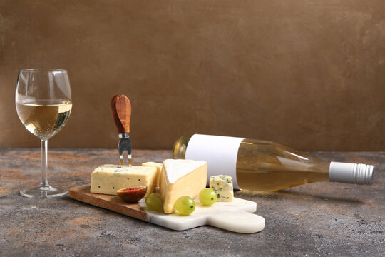 Board with tasty cheese, grapes, fig, bottle and glass of wine on table near brown wall