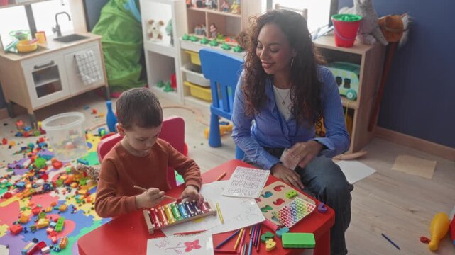 Boy plays xylophone at a small red table while teacher raises hand and smiles in a preschool building strewn with toys and art supplies; learning support joy.