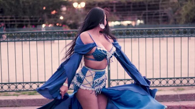 Sensual Latina woman dancing belly dance, twirling in slow motion, wearing a beautiful blue dress, in a park at sunset, with makeup. Dancing and smiling, with sensuality and joy.