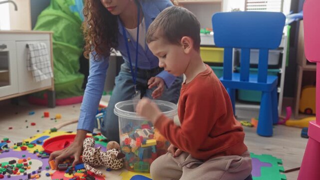 Boy playing with colorful blocks and toys while teacher kneels and guides play near a toy kitchen and blue chair in a building; learning calm.