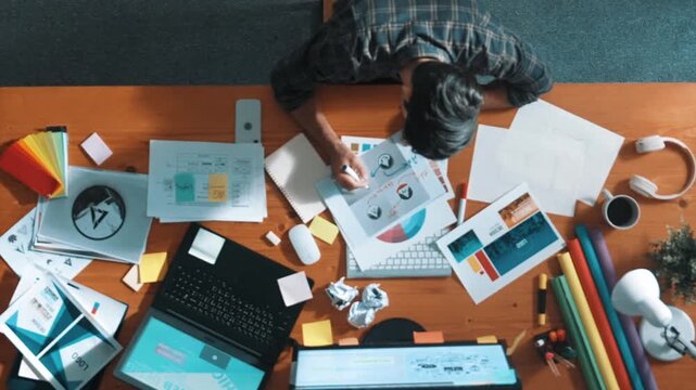 Top view of designer writing and designing logo graphic at meeting. Closeup of business man working on colored palettes and writing idea on sticky notes on table with equipment and paper. Symposium.
