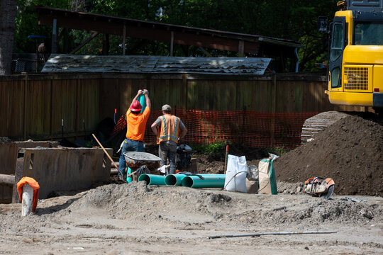 Stormwater system pipe installation in progress, construction workers with 6 in green PVC pipes standing above excavated dirt trench on infrastructure work site
