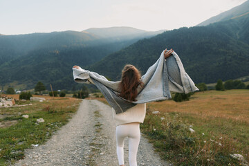 Fototapeta premium A young woman walks along a rural path through a sunlit valley, arms raised as a knitted shawl billows behind her, expressing freedom and outdoor lifestyle.