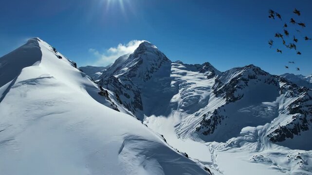 Panoramic aerial view of high altitude snow-covered mountain peaks under a clear blue sky with bright sunlight and lens flare, featuring sharp ridges, rocky outcrops, and icy slopes.