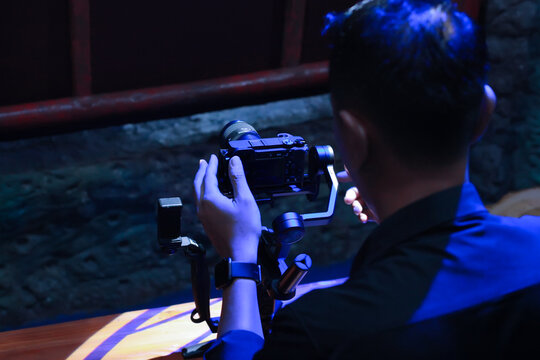 medium shot of a man operating a mirrorless camera mounted on a 3-axis handheld gimbal stabilizer in a dimly lit studio environment