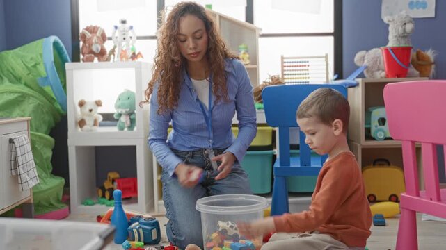 Woman teacher kneeling, raising open hand for high five while boy sorts colorful toys into a plastic bucket in a childcare building; encouragement bonding.