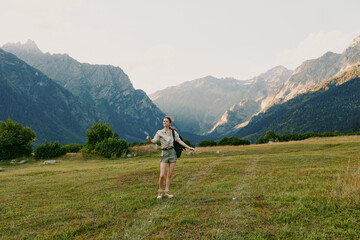 Fototapeta premium Young woman with long hair walks across a green meadow surrounded by mountains during a sunny day, enjoying nature and outdoor adventure with a casual outfit.