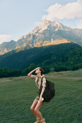 Fototapeta premium Cheerful woman outdoors, hiking in a sunlit meadow beneath mountains, with a backpack, relaxed posture, natural colors, fresh breeze, adventurous vibe with joyful energy