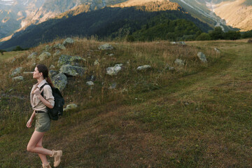 Naklejka premium Cheerful woman hiking outdoors with backpack, casual attire, grassy trail, mountain ridge in background, natural light, energetic lifestyle, scenic nature moment outdoors