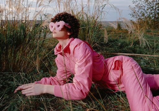 Pink fashion model lying in tall grass outdoors, wearing pink sunglasses and a matching pink suit, dramatic pose, natural light, editorial style in nature, bold mood, cinematic