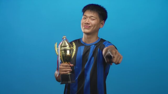 Man holding trophy and points finger while wearing a blue soccer uniform in studio; pride victory celebration.