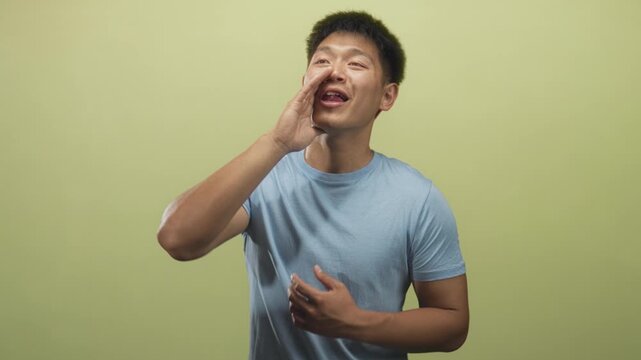 Man cupping hand to mouth shouting into space in studio while wearing blue t shirt and gesturing with other hand; playful call.