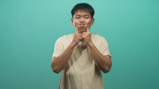 Man pressing his cheeks with index fingers and puffing lips while posing against a teal studio wall, visible forearms and shirt; playful.