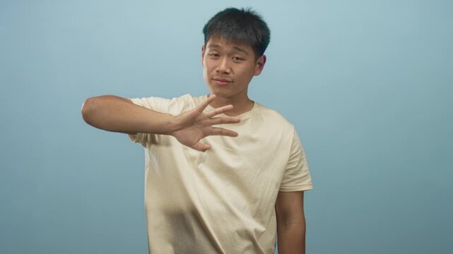 Young chinese man in beige t shirt waves hand with visible forearm toward camera against light blue studio wall; friendly greeting.