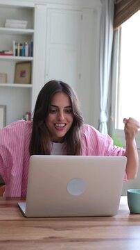 A happy young woman shows extreme joy and celebrates a great achievement while working on her laptop in her home office. Her reaction captures the triumph of a successful online endeavor.