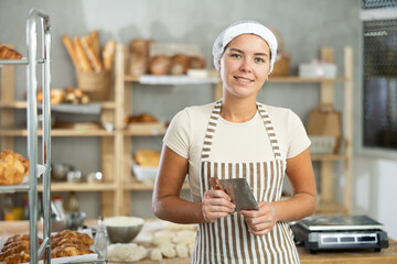 While working with dough and making buns, baker girl takes break, stands resting. Near cutting table in kitchen, employee prepares dough mass for making croissants. © JackF