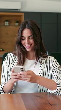 A cheerful young woman in a striped shirt happily uses her smartphone, engaging in digital communication. She occasionally looks away thoughtfully, enjoying a moment of online connection or reflecti