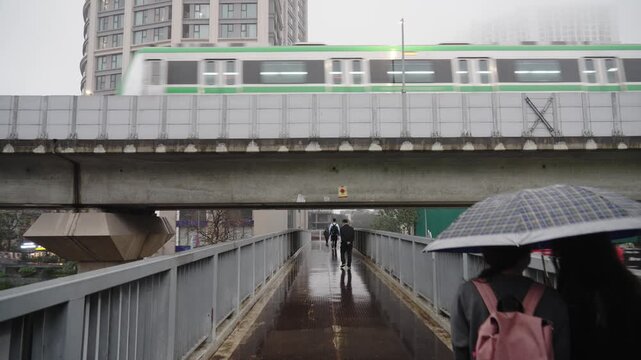 A metro train passes above Nguyen Trai street while pedestrians walk below with umbrellas on a rainy day in Hanoi city.