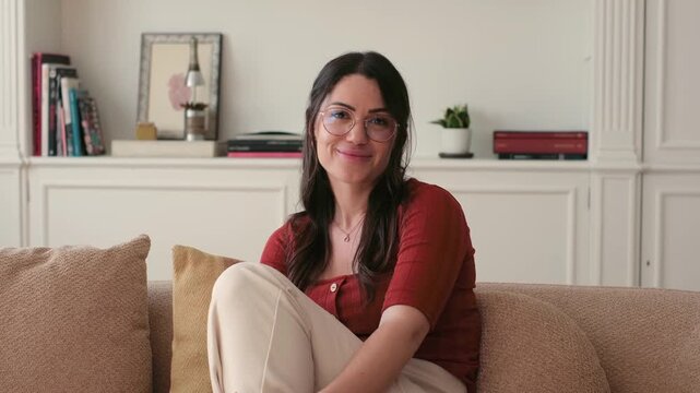 A joyful woman in glasses smiles genuinely while relaxing on a comfortable sofa at home. She exudes happiness and a sense of calm in her cozy living space.