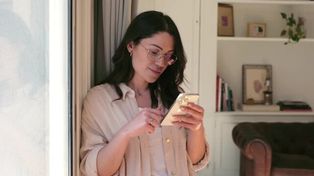 A relaxed young woman in glasses happily scrolls through her smartphone, enjoying digital communication and browsing online content in the comfort of her modern home. Natural daylight streams throug