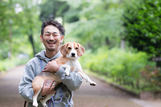 新緑背景で愛犬のビーグルを抱く男性の笑顔、公園で過ごす幸せなお散歩時間のカメラ目線クローズアップ