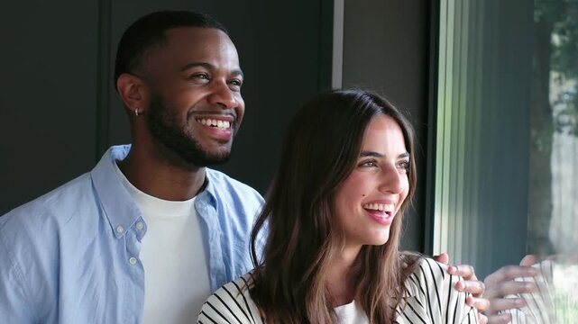 A happy multiethnic couple smiles joyfully and shares a loving moment as they look out a window, perhaps reflecting on their future or enjoying a peaceful day at home.