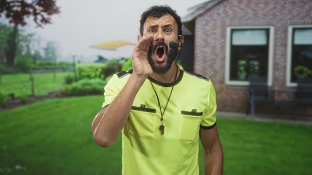 Man referee with whistle cupping hand to mouth shouting on residential building lawn wearing yellow shirt; authority urgent call.