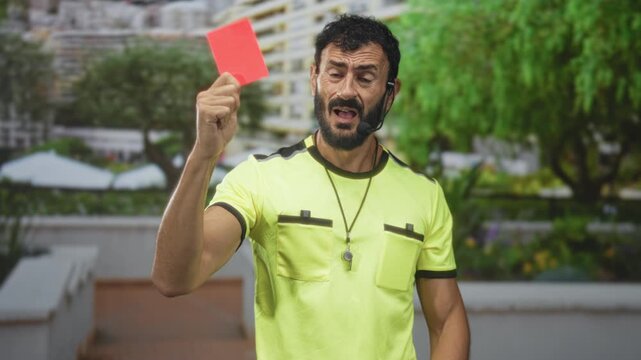 Referee man holding red card and whistle, headset mic attached, hand raised in building courtyard with trees and terraces; authority enforcement.