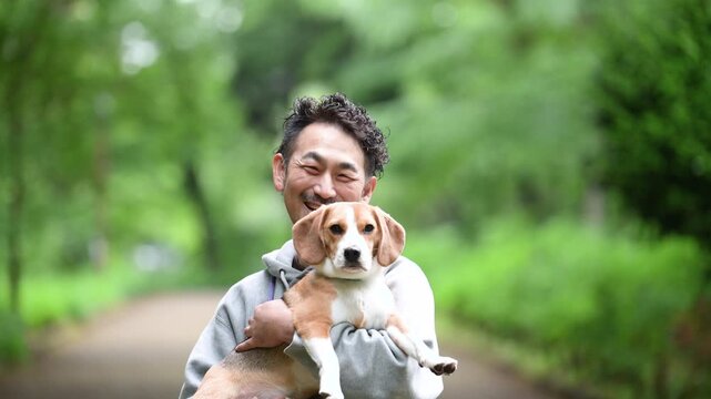 新緑背景で愛犬のビーグルを抱く男性の笑顔、公園で過ごす幸せなお散歩時間のカメラ目線クローズアップ動画	