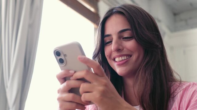 A joyful young woman smiles and laughs while scrolling on her mobile phone, enjoying content or communicating with friends in a bright, relaxed home setting.