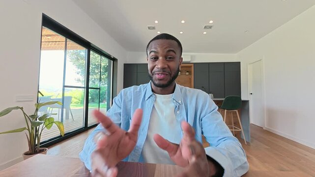 A young African American man smiles and gestures enthusiastically during a video call from his bright, modern home. He appears engaged and happy, possibly sharing ideas or having a friendly conversa