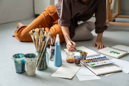 Creative process at home art studio, talented woman painter drawing picture sitting on floor surrounded by art supplies, professional female artist creating painting on paper with watercolors.