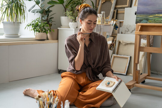 Positive talented woman artist enjoying finished watercolor sketch in notebook, looking at picture adding final touches, smiling female painter practicing working with aquarelle media in sketchbook.