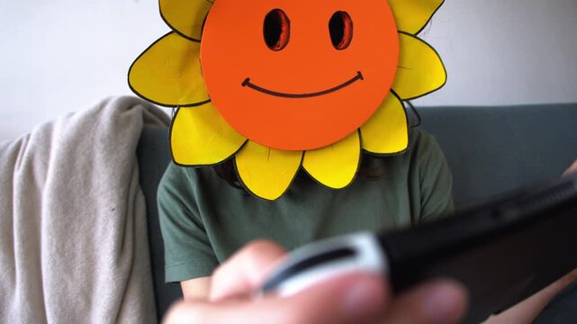 A masked teenager plays a video game on his console while sitting on the couch wearing a sunflower mask and remaining incognito. A boy plays a video game. An incognito player.