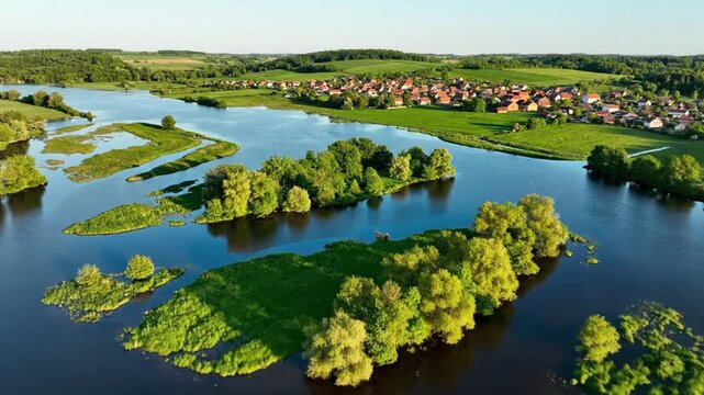 Panoramic aerial view of a flooded landscape featuring lush green islands of trees and grasslands, with a remote village and rolling hills in the background on a sunny day.