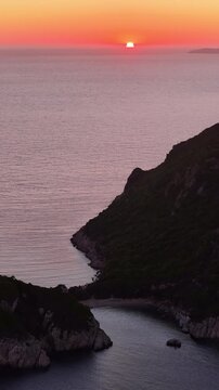 Vertical aerial shot of a magnificent red sunset dipping into the Ionian sea horizon next to the dark silhouette cliffs of Afionas, Corfu, Greece