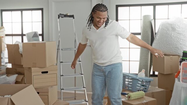 Man spreads arms amid stacked cardboard moving boxes, ladder, laundry basket and rolled rug inside a spacious building room with packing materials; fresh start joy.