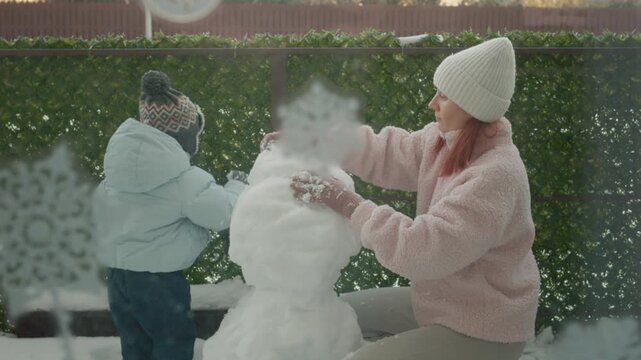 A mother and her young son gather snow to make a snowman in the yard outside their apartment. They enjoy their time together as they shape and decorate the snowman.