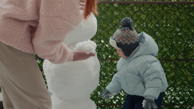 A mother and her young son spend time together building a snowman in the yard of their apartment building. They have fun while playing in the snow.
