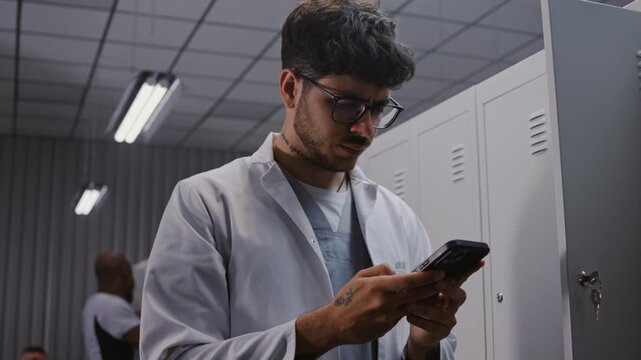 Young Male Physician in White Coat Stands by Locker After Shift, Intensely Reviewing Urgent Laboratory Results and Patient Diagnostics on Smartphone. Concept of Medical Urgency and Dedication.