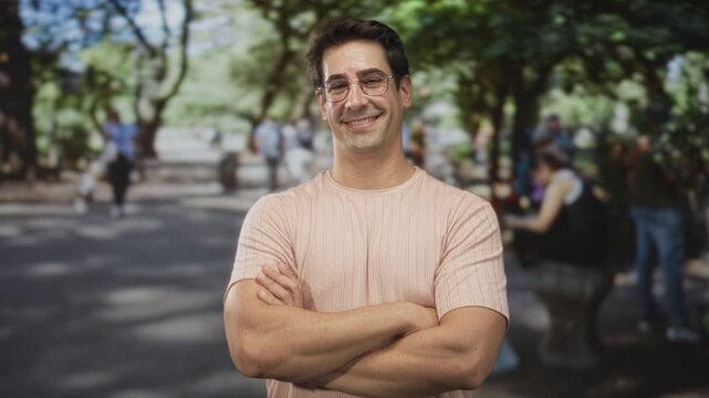 Man smiling with arms crossed and right hand giving thumbs up, wearing pink t shirt and glasses in street park with benches and trees visible; contentment.